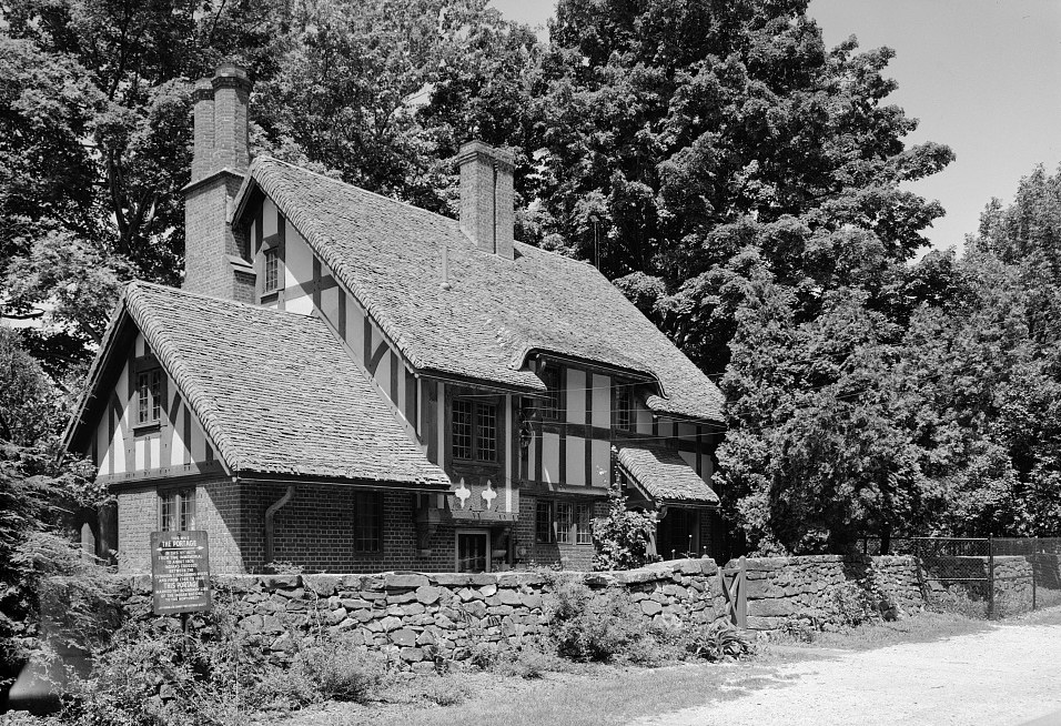 A Tudor-style cottage with steep shingled roofs, half-timbered upper walls, brick below and two tall chimneys, behind a low fieldstone wall under tall trees.