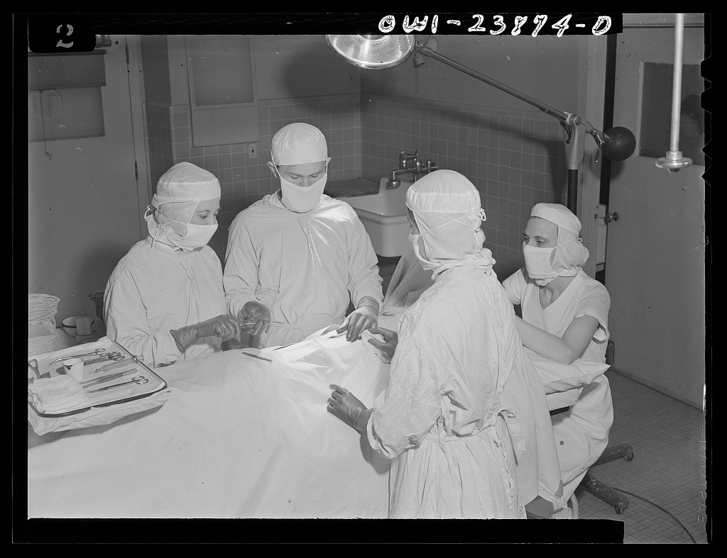 A black-and-white photograph of a surgeon at work in an operating room, with a student nurse handing him instruments.