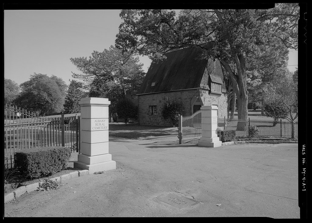 The stone and iron south entrance of a large rural cemetery, with trees beyond.