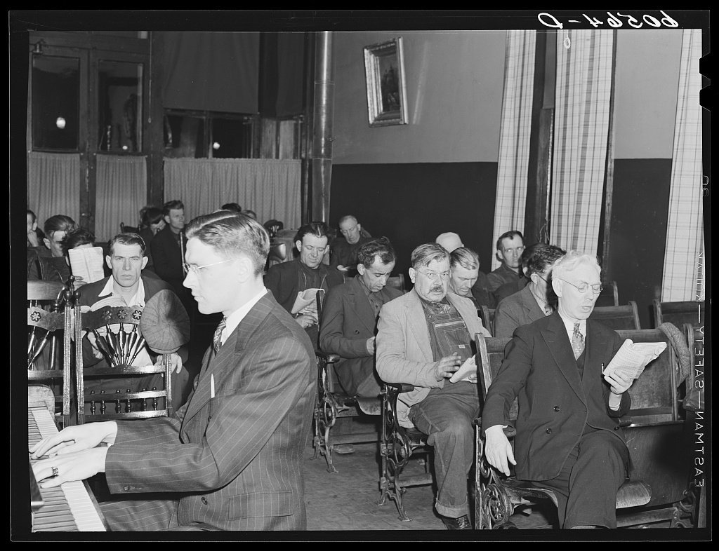 Men seated on benches in a mission hall, singing from hymnbooks.