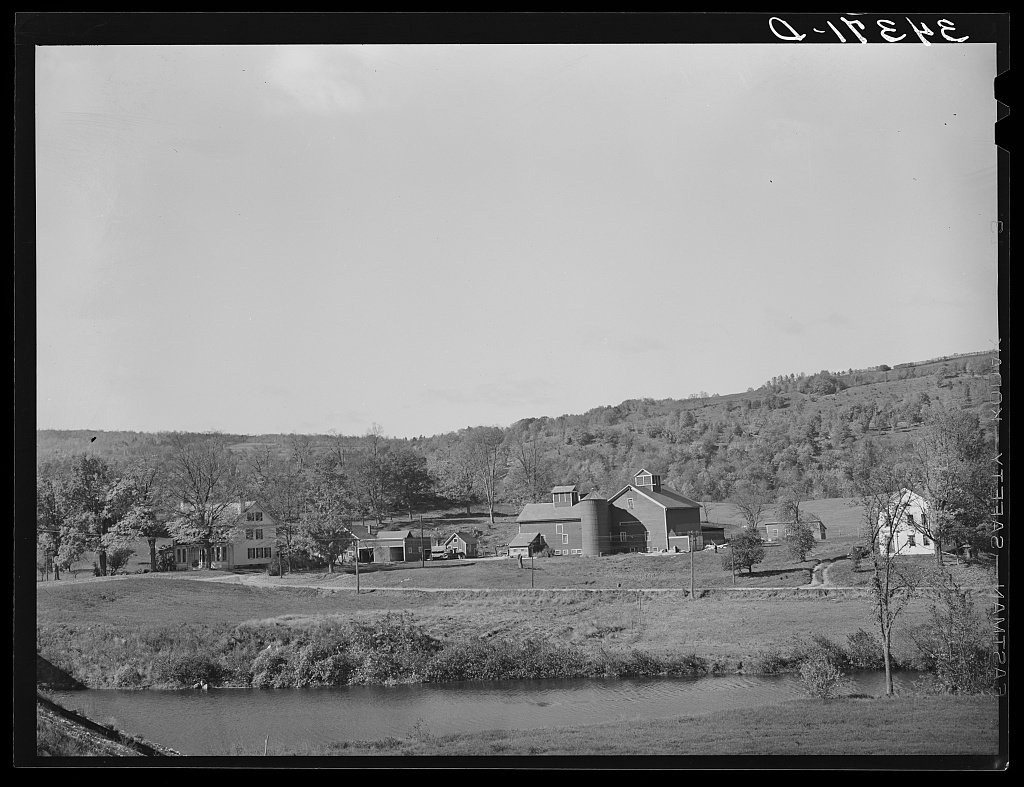A white farmhouse and barns beside a road, with wooded hills behind.